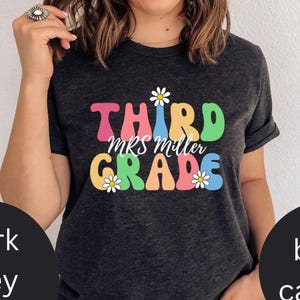 May include: Dark grey t-shirt featuring the text "THIRD MRS MILLER GRADE" in a retro font with daisy accents. The letters are in various bright colours. A casual, everyday tee.