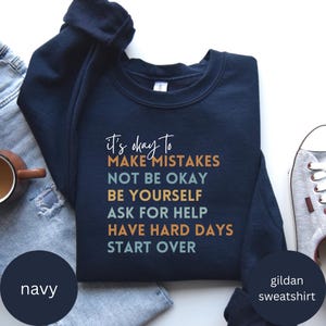 Peut inclure: Sweat-shirt bleu marine avec le texte "it's okay to MAKE MISTAKES NOT BE OKAY BE YOURSELF ASK FOR HELP HAVE HARD DAYS START OVER" imprimé en orange, bleu et jaune.