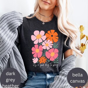 May include: Dark grey heather t-shirt with a floral design in pink, orange, and white. The shirt features the text "It's a good day to play" in a handwritten style. The shirt is made of Bella Canvas material.