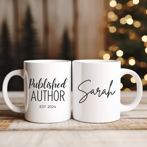 May include: Two white ceramic mugs. One mug says "Published Author Est. 2024" and the other mug says "Sarah" in cursive.