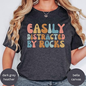 Könnte beinhalten: Dunkelgraues Heather-T-Shirt mit dem Text "EASILY DISTRACTED BY ROCKS" in einer farbenfrohen Retro-Schriftart. Das Shirt besteht aus Bella Canvas-Material.