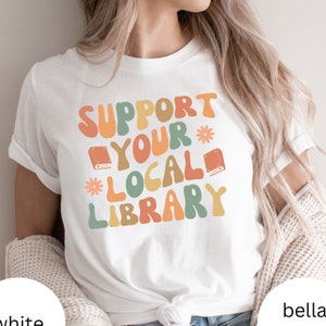 Könnte beinhalten: Weißes T-Shirt mit einer Grafik im Retro-Stil, auf dem "Unterstütze deine lokale Bibliothek" steht, mit Illustrationen von Büchern und Blumen.