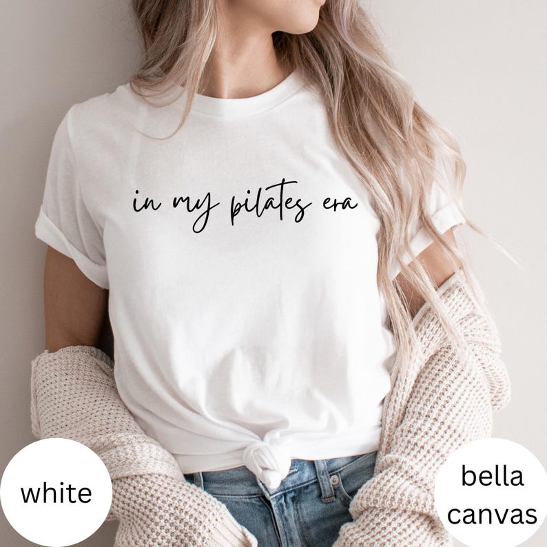 Pu&ograve; includere: T-shirt bianco con la scritta "in my pilates era" in stile scritto a mano. La maglietta &egrave; del marchio Bella + Canvas. La modella indossa jeans blu e un maglione color crema.