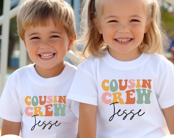 Può includere: Due bambini sorridenti indossano magliette bianche. Ogni maglietta presenta la scritta "COUSIN CREW" in lettere retrò colorate, con il nome "Jesse" in corsivo.