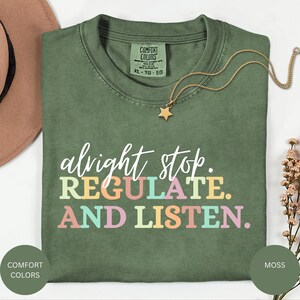 Puede incluir: Una camiseta verde musgo con el texto "alright stop. REGULATE. AND LISTEN." en colores pastel. Un collar dorado con un colgante de estrella descansa sobre la camiseta. La camiseta tiene una etiqueta Comfort Colors.