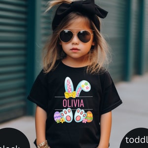 May include: Black t-shirt featuring a colourful Easter bunny design with the name "Olivia" printed on it. The bunny has blue ears with pink polka dots and a yellow bow. The bunny's feet are blue with white paw prints. The shirt also includes colourful Easter eggs and flowers.