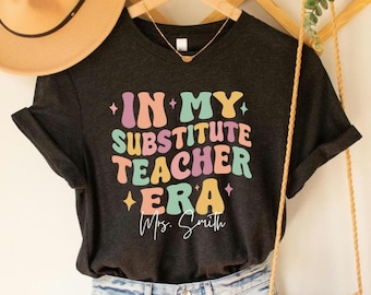 Könnte beinhalten: Ein dunkelgraues T-Shirt mit dem Text "In My Substitute Teacher Era" in bunten, Retro-Lettern. Der Text ist von kleinen Sternen umgeben. Der Name "Mrs. Smith" ist darunter in Kursivschrift geschrieben.