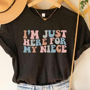 Puede incluir: Una camiseta gris oscuro con el texto "I'm Just Here For My Niece" en letras retro de colores.
