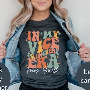 May include: A dark grey t-shirt with the text "In My Vice Principal Era" in colourful retro-style lettering. The text "Mrs. Smith" is printed below the main text.