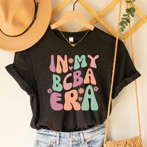 Pode incluir: Uma camiseta cinza escura com o texto "In My BCBA Era" em letras coloridas de estilo retrô. As letras são em tons de rosa, verde, laranja e roxo.