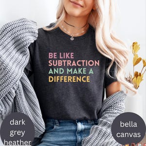 May include: Dark grey heather t-shirt with the text "BE LIKE SUBTRACTION AND MAKE A DIFFERENCE" in pink, green, and yellow. The shirt is made of Bella Canvas material.