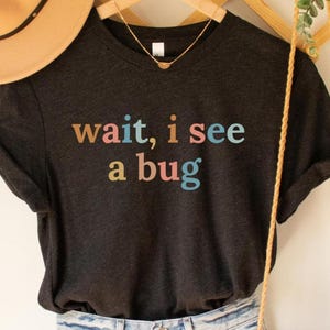May include: Dark grey t-shirt with the phrase "wait, i see a bug" in colourful, retro-style lettering. The shirt is hanging on a wooden hanger, with a gold necklace visible.