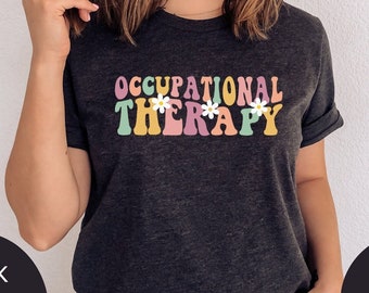 Op de afbeelding: Een donkergrijs T-shirt met de tekst "OCCUPATIONAL THERAPY" in kleurrijk retro-lettertype met witte madeliefjes.