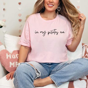Pu&ograve; includere: Una maglietta rosa chiaro con la scritta "in my pilates era" in corsivo nero. La maglietta &egrave; abbinata a jeans blu. L'immagine mostra una persona seduta su un divano bianco.