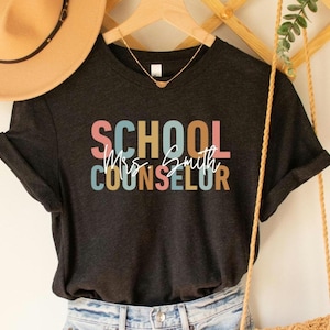 May include: Dark gray t-shirt with the words "SCHOOL COUNSELOR Mrs. Smith" in colorful block letters and cursive script. The shirt is hanging on a wooden hanger with a gold necklace and a straw hat in the background.