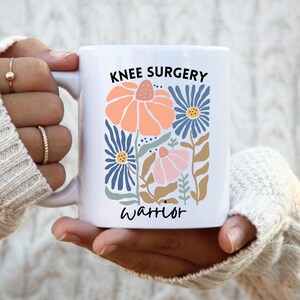 Könnte beinhalten: Weiße Keramik-Tasse mit den Worten "KNEE SURGERY" und "warrior" in schwarzer Schrift. Die Tasse zeigt ein Blumendesign in Orange-, Blau-, Rosa- und Brauntönen. Die Tasse wird von einer Person gehalten, die einen weißen Pullover trägt.