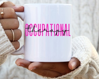 May include: A white ceramic mug featuring the words "OCCUPATIONAL THERAPY" in bright pink block letters and a black cursive font. The mug is held by a person wearing a cream-coloured sweater.