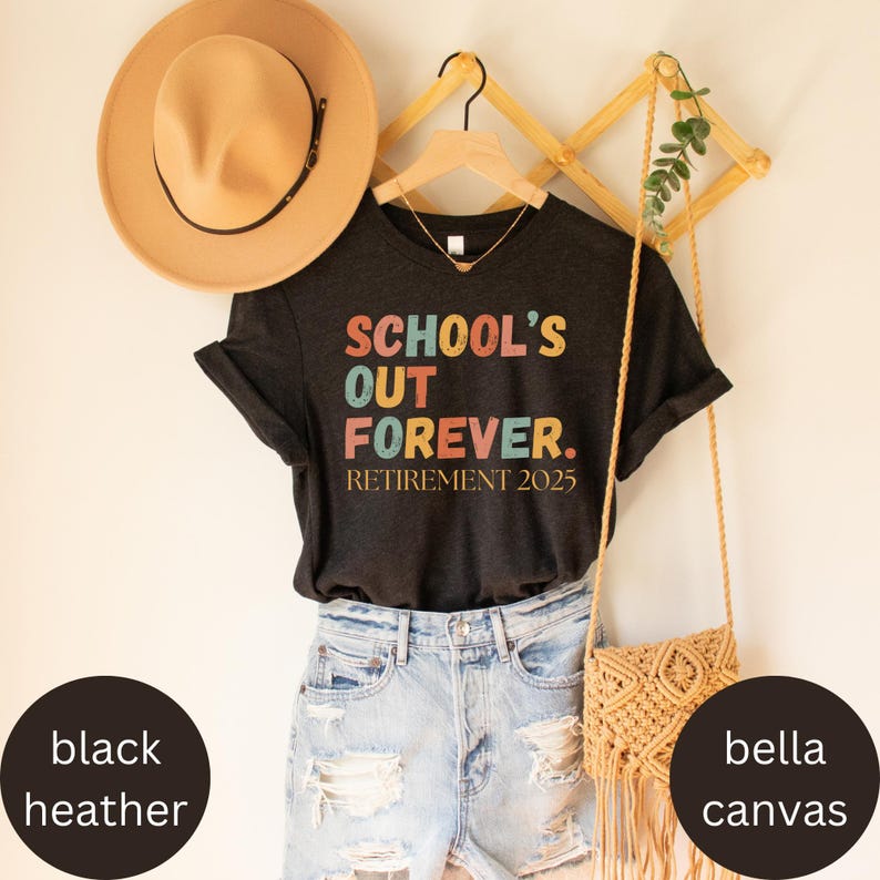May include: A black heather v-neck t-shirt with the text "SCHOOL'S OUT FOREVER. RETIREMENT 2025" in colorful letters. The shirt is paired with denim shorts, a tan hat, and a woven crossbody bag. The shirt is hanging on a wooden hanger.