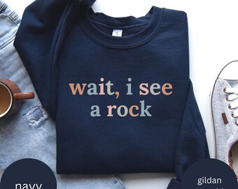 Puede incluir: Una sudadera Gildan azul marino con el texto "wait, i see a rock" impreso en colores pastel.