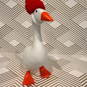 May include: A white goose figurine with a red knit hat and orange feet and beak. The goose is wearing a red knit hat with a white pom-pom. The goose is standing on a surface with a geometric pattern.