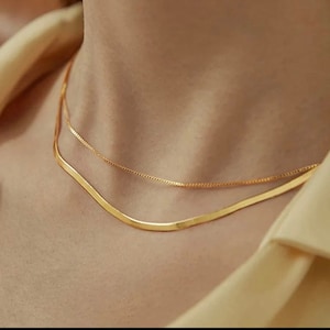 May include: Two gold necklaces layered on a person's neck. The top necklace is a delicate chain, while the bottom necklace is a thicker, flat chain.