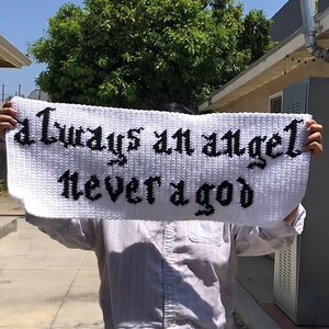 Op de afbeelding: Witte gehaakte banner met zwarte tekst die "always an angel never a god" luidt.
