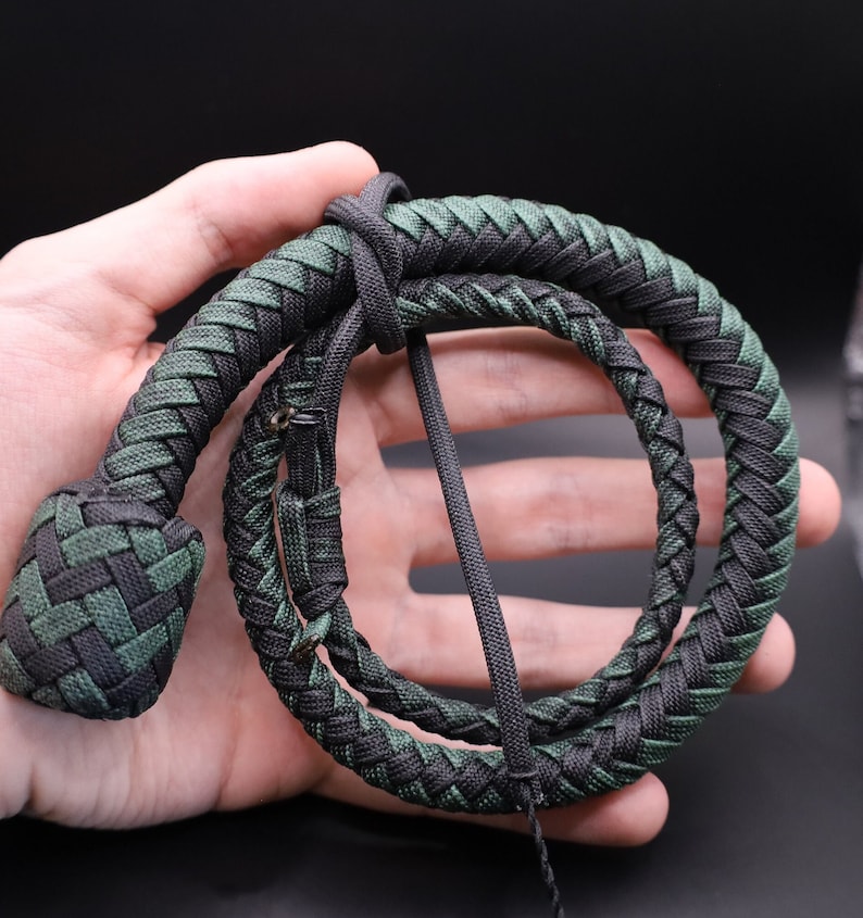 May include: A braided whip in shades of green and black, coiled in a loop. The whip's handle is a tightly woven knot, and the body is a series of interwoven strands. The overall design is symmetrical and detailed.
