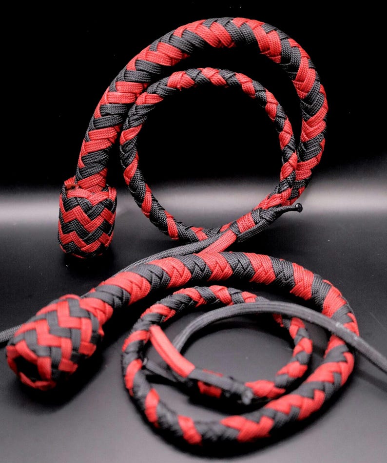 May include: A braided whip in red and black. The whip is made of interwoven cords, with a rounded handle and a long, flexible body. The design features a repeating pattern of red and black, creating a visually striking effect.