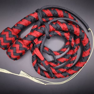 May include: A braided whip in red and black. The whip is made of interwoven cords, with a handle and a long, flexible body. The handle is a solid, tightly woven braid. The whip's body is a series of braided loops.
