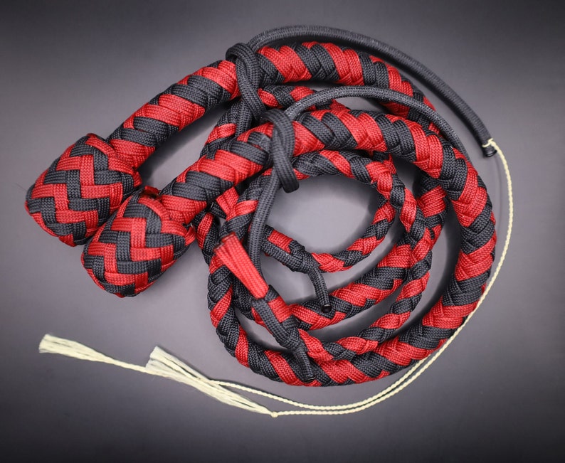 May include: A braided whip in red and black. The whip is made of interwoven cords, with a handle and a long, flexible body. The handle is a solid, tightly woven braid. The whip's body is a series of braided loops.
