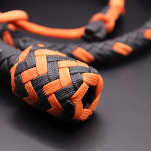 May include: Close-up of a braided rope in black and orange. The rope is tightly woven, with a prominent, rounded knot at the end. The color scheme is a striking contrast of black and bright orange, creating a visually appealing texture.