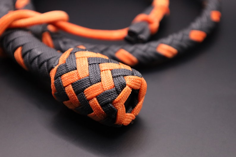 May include: Close-up of a braided rope in black and orange. The rope is tightly woven, with a prominent, rounded knot at the end. The color scheme is a striking contrast of black and bright orange, creating a visually appealing texture.