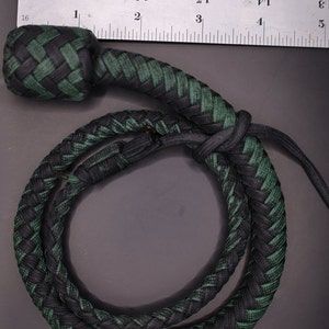 May include: A braided whip in black and green, with a rounded knot at the top. The whip is measured against a ruler, showing its length. The braid is tightly woven, creating a durable and textured appearance.