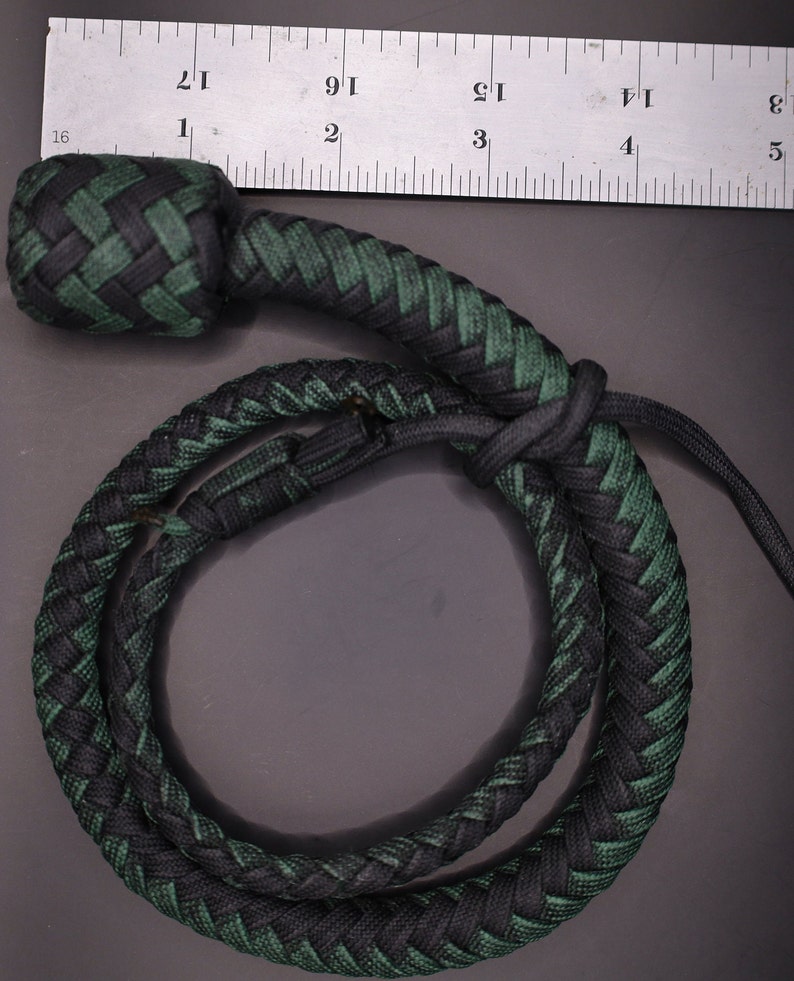 May include: A braided whip in black and green, with a rounded knot at the top. The whip is measured against a ruler, showing its length. The braid is tightly woven, creating a durable and textured appearance.