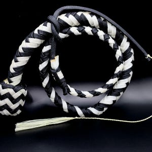 May include: A braided whip in black and white. The whip is made of interwoven strands, with a rounded handle and a long, tapering body. A tassel hangs from the end. The background is black.
