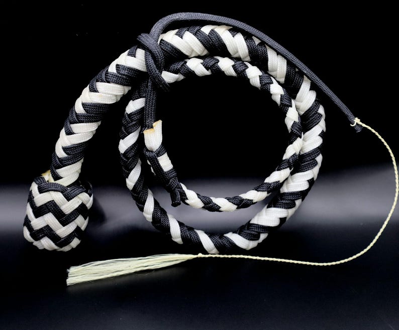 May include: A braided whip in black and white. The whip is made of interwoven strands, with a rounded handle and a long, tapering body. A tassel hangs from the end. The background is black.
