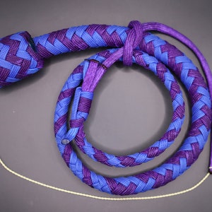 May include: A braided whip in shades of blue and purple, with a rounded handle and a long, spiraling body. The whip's design features a repeating pattern of interwoven strands. A thin, off-white cord with multiple strands extends from the end.