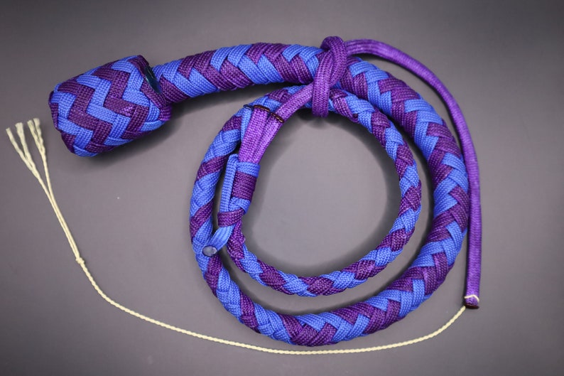 May include: A braided whip in shades of blue and purple, with a rounded handle and a long, spiraling body. The whip's design features a repeating pattern of interwoven strands. A thin, off-white cord with multiple strands extends from the end.