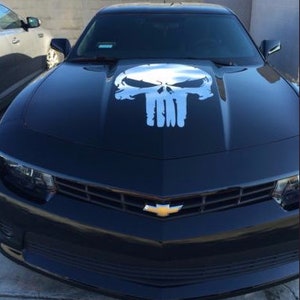 May include: Black sports car featuring a white skull graphic on the bonnet. The car has a black grille and a gold Chevrolet emblem. The car is parked outdoors.