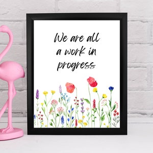 May include: A framed print with a white background and black frame. The print features a watercolor illustration of wildflowers and the text "We are all a work in progress".
