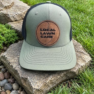 May include: A green and black trucker cap with a leather patch. The patch is round and features the text "LOCAL LAWN CARE". The cap is resting on a stone. The cap has a curved peak.