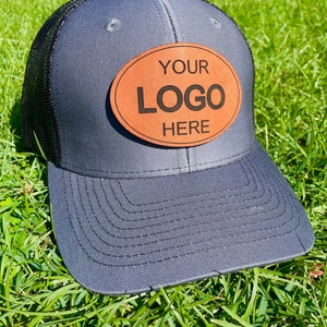 May include: A grey and black mesh trucker hat with a brown oval leather patch that says "YOUR LOGO HERE".