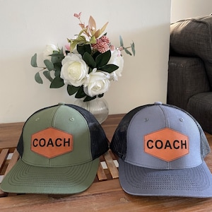 May include: Two baseball caps with black mesh backs and solid front panels. One cap is olive green and the other is slate blue. Each features a hexagonal tan leather patch on the front centre with the word COACH embossed in black capital letters.