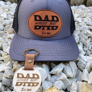 May include: A grey trucker hat with a black mesh back and a tan oval leather patch featuring the text DAD WORLD'S BEST DAD Est. 2019. A matching round wooden keyring with the same text and date is attached to the brim of the hat.