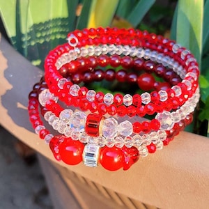 May include: A set of five beaded bracelets in red and clear colors. The bracelets are stacked on top of each other and are made of small, round beads.