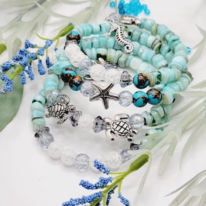 Può includere: Un set di cinque braccialetti con ciondoli a forma di cavalluccio marino, stella marina e tartaruga. I braccialetti sono realizzati con perline turchesi e bianche.