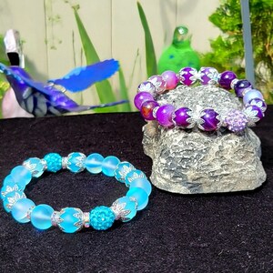 Op de afbeelding: Twee armbanden met zilveren accenten en gekleurde kralen. De ene armband heeft lichtblauwe kralen met een turquoise middelste kraal. De andere armband heeft paarse en roze agaat kralen met een paarse middelste kraal.
