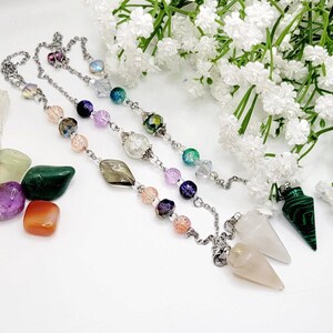 May include: Three silver chains with crystal beads and pendants. The pendants are a white quartz point, a green malachite point, and a clear crystal point. The chains are on a white background with small green and purple stones.