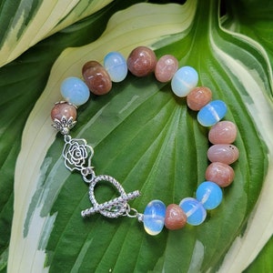 Könnte beinhalten: Ein Armband aus rosafarbenen und weißen Edelsteinperlen mit einem silbernen Verschluss mit Blumenmuster.