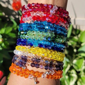 May include: A stack of colorful beaded bracelets in shades of red, blue, green, yellow, purple, and orange. Each bracelet is made of small, faceted beads. Some bracelets have small charms.